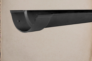 A close-up product shot of a black metal gutter section mounted against a large beige wall. The words 'TRADCAST by Guttercrest' appear in the upper right corner.